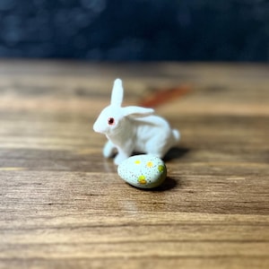 May include: A white bunny figurine with red eyes sits near a speckled Easter egg. The egg is light green with yellow and white floral accents. The bunny and egg are on a wooden surface.