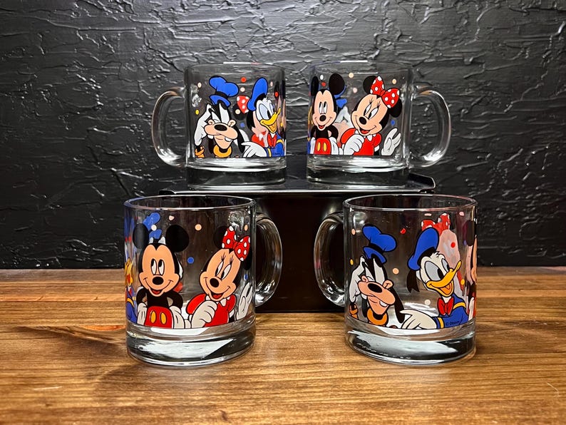 May include: Set of four clear glass mugs featuring Disney characters Mickey Mouse, Minnie Mouse, Donald Duck, and Goofy. Each mug has a handle and is decorated with colorful cartoon images. The mugs are displayed on a wooden surface.