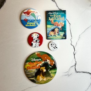 May include: Five Disney-themed button pins featuring characters from The Fox and the Hound and 101 Dalmatians. The pins are round and have a white background with colorful images of the characters. One pin features a red background with a Dalmatian dog and puppies. One pin features a blue background with a mermaid and a fish. One pin features a white background with a fox and a hound dog. One pin features a white background with a Dalmatian dog. One pin features a white background with a fox and a hound dog.