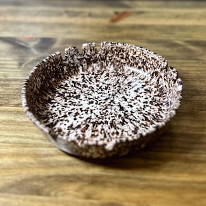 May include: A speckled ceramic ashtray with a brown and white pattern. The ashtray has a round shape with notched edges for resting cigarettes. The interior is white with brown speckles, and the rim is brown.
