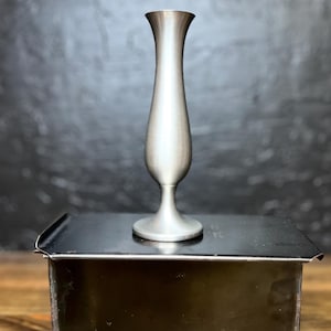 May include: A brushed metal vase with a slender, elongated body and a flared top. The vase is silver in color and stands on a round base. It is displayed on a black box, with a dark background.