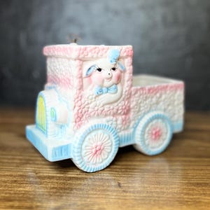 May include: A pink and blue ceramic planter shaped like a pickup truck with a white sheep face on the front. The truck has a blue bow tie and a pink and blue floral pattern.
