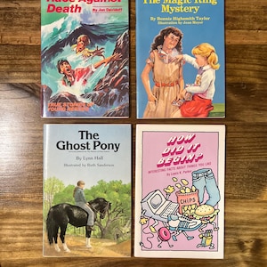 May include: Four vintage children's books are displayed on a wooden surface. The titles include "Race Against Death," "The Magic Ring Mystery," "The Ghost Pony," and "How Did It Begin?" Each book features colorful illustrations and text.