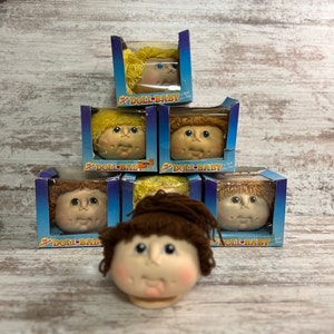 May include: A stack of six doll head kits in blue boxes with the text "The Original Doll-Baby" on the front. The doll heads have brown and blonde yarn hair and are in various stages of completion.