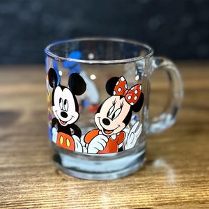 May include: Clear glass mug featuring Mickey Mouse and Minnie Mouse. Mickey is wearing red shorts and white gloves. Minnie is wearing a red dress and a red bow with white polka dots. The mug has a handle and is on a wooden surface.