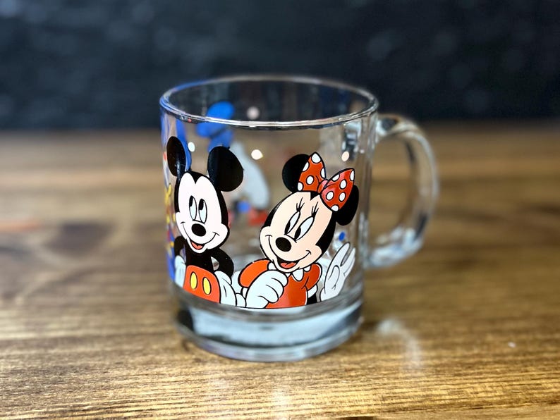 May include: Clear glass mug featuring Mickey Mouse and Minnie Mouse. Mickey is wearing red shorts and white gloves. Minnie is wearing a red dress and a red bow with white polka dots. The mug has a handle and is on a wooden surface.