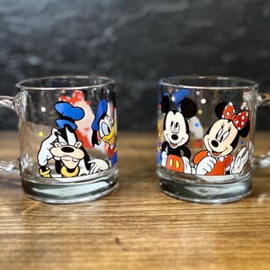 May include: Two clear glass mugs with cartoon character designs. One mug features Goofy, Donald Duck, and another character. The second mug showcases Mickey and Minnie Mouse. Both mugs have a handle and a rounded base.