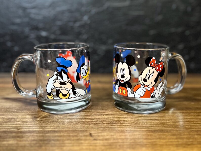 May include: Two clear glass mugs with cartoon character designs. One mug features Goofy, Donald Duck, and another character. The second mug showcases Mickey and Minnie Mouse. Both mugs have a handle and a rounded base.