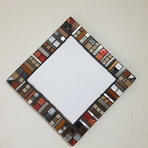 Könnte beinhalten: Ein rautenförmiger Spiegel mit einem Mosaikrahmen. Der Rahmen besteht aus kleinen, bunten Fliesen in Braun-, Orange- und Silbertönen.