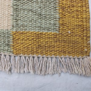 May include: Close-up of a woven rug with a geometric pattern. The rug features squares of cream, sage green, and mustard yellow. The bottom edge has a fringe of cream-colored yarn.