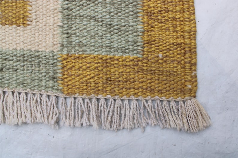 May include: Close-up of a woven rug with a geometric pattern. The rug features squares of cream, sage green, and mustard yellow. The bottom edge has a fringe of cream-colored yarn.