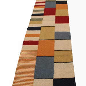 May include: A rectangular, multi-colored runner rug with a patchwork design. The rug features blocks of red, orange, beige, blue, and black. The rug also has striped sections in orange and beige. The rug is made of woven material.