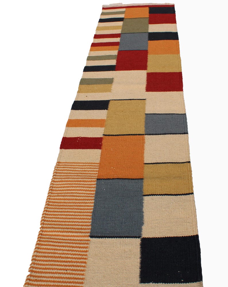 May include: A rectangular, multi-colored runner rug with a patchwork design. The rug features blocks of red, orange, beige, blue, and black. The rug also has striped sections in orange and beige. The rug is made of woven material.