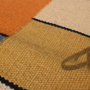 May include: Close-up of a woven rug with a patchwork design. The rug features sections of orange, tan, yellow, and blue, separated by thin black lines. The texture of the rug is visible, showing the individual fibers.