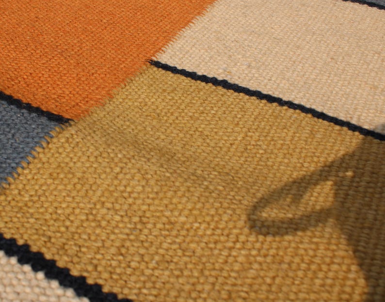 May include: Close-up of a woven rug with a patchwork design. The rug features sections of orange, tan, yellow, and blue, separated by thin black lines. The texture of the rug is visible, showing the individual fibers.