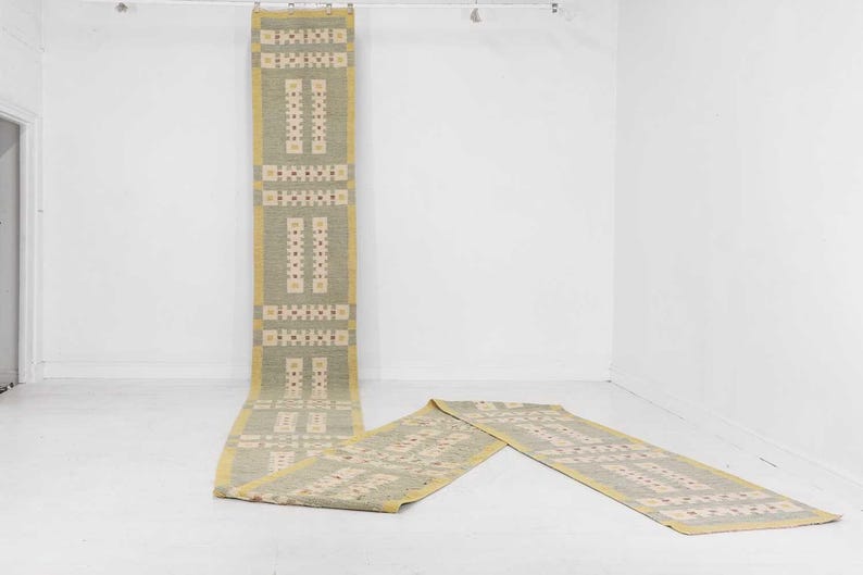 May include: A long, rectangular rug with a repeating geometric pattern in beige, olive green, and yellow. The rug is hanging from a metal bar and also lying on the white floor. The rug's design features rectangular and square shapes.