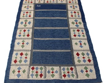 May include: A rectangular blue rug with a cream border featuring repeating geometric patterns in red, green, and blue. The rug has a central blue field with horizontal stripes and the name "Ava" in the upper center.