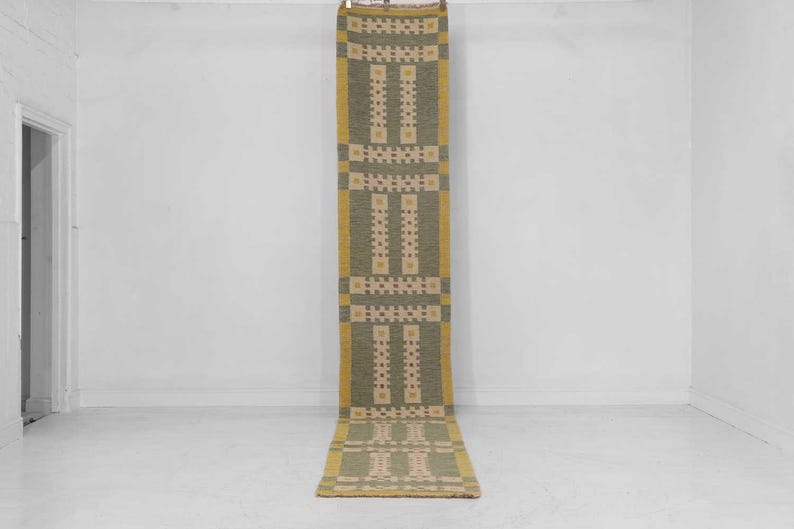 May include: A long, rectangular rug with a repeating geometric pattern. The rug features a central olive green field with cream and yellow accents. The edges are bordered with a yellow hue. The rug is hanging vertically against a white wall.