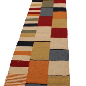 May include: A rectangular, multi-colored rug with a patchwork design. The rug features blocks of red, orange, beige, blue, and black. The rug is made of woven material and has a striped pattern on one end.