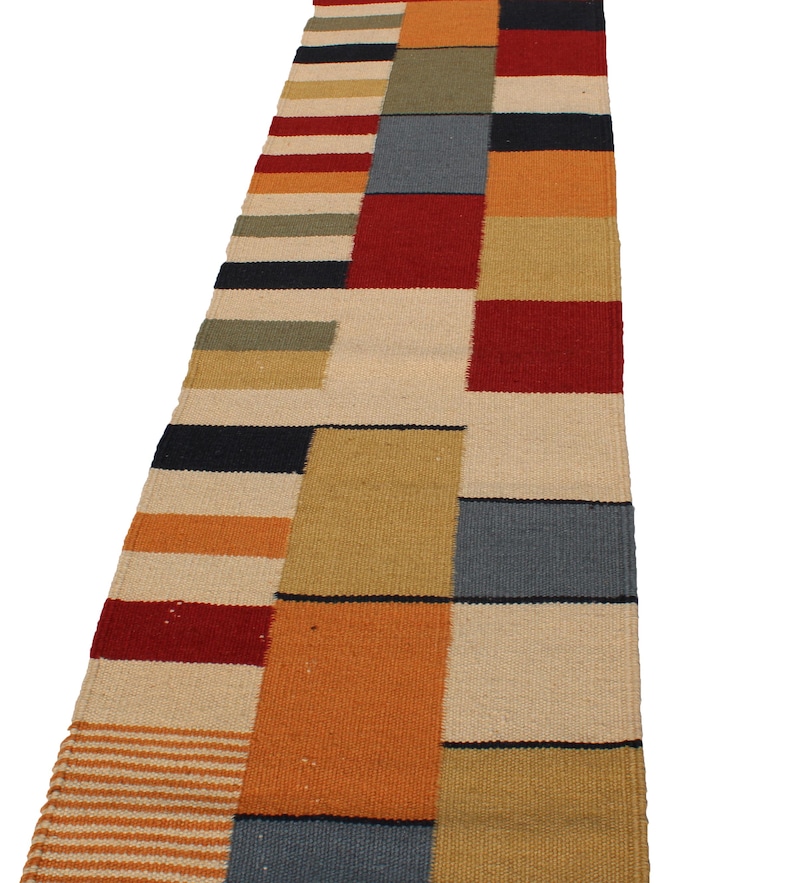 May include: A rectangular, multi-colored rug with a patchwork design. The rug features blocks of red, orange, beige, blue, and black. The rug is made of woven material and has a striped pattern on one end.