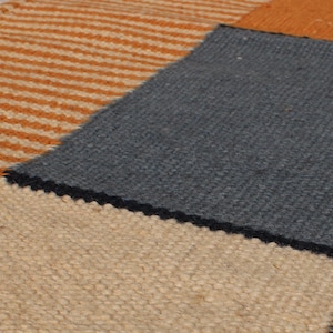 May include: Close-up of a patchwork rug with various textured sections. The rug features a striped orange and cream section, a solid gray square, and other squares in orange, cream, black, and yellow. The rug has a woven texture.