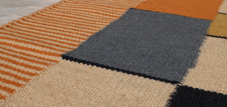 May include: Close-up of a patchwork rug with various textured sections. The rug features a striped orange and cream section, a solid gray square, and other squares in orange, cream, black, and yellow. The rug has a woven texture.