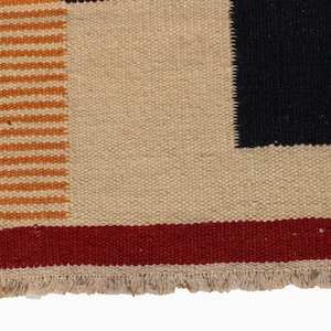 May include: Close-up of a woven textile with a geometric design. The rug features a beige background with a black square, a red stripe, and orange horizontal stripes. The bottom edge has a fringe.