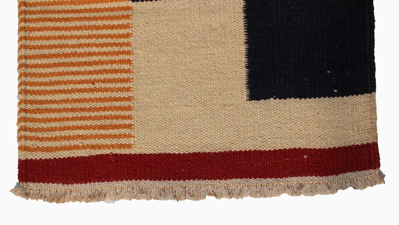 May include: Close-up of a woven textile with a geometric design. The rug features a beige background with a black square, a red stripe, and orange horizontal stripes. The bottom edge has a fringe.