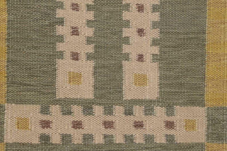 May include: Close-up of a woven textile with a geometric pattern. The design features repeating shapes in off-white, olive green, and yellow. The texture appears to be coarse and durable, suitable for a rug or upholstery.