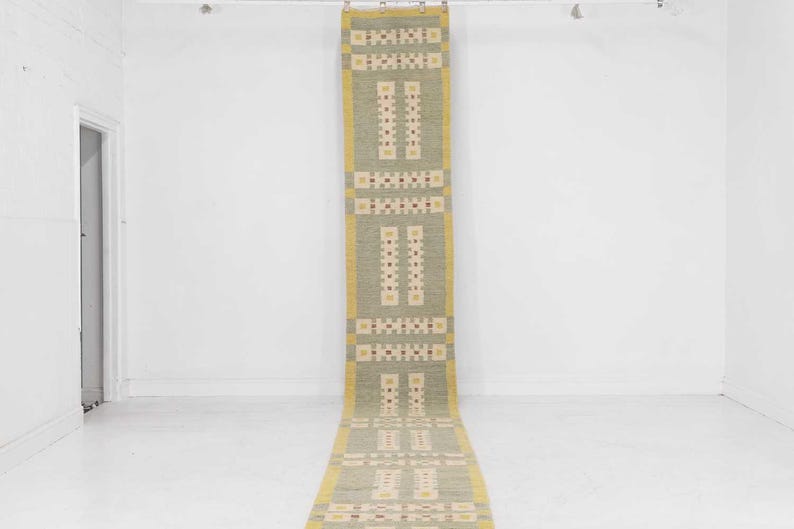May include: A long, rectangular rug with a repeating geometric pattern. The rug features a light green background with cream and red accents. The edges are bordered with a yellow trim. The rug is hanging vertically.
