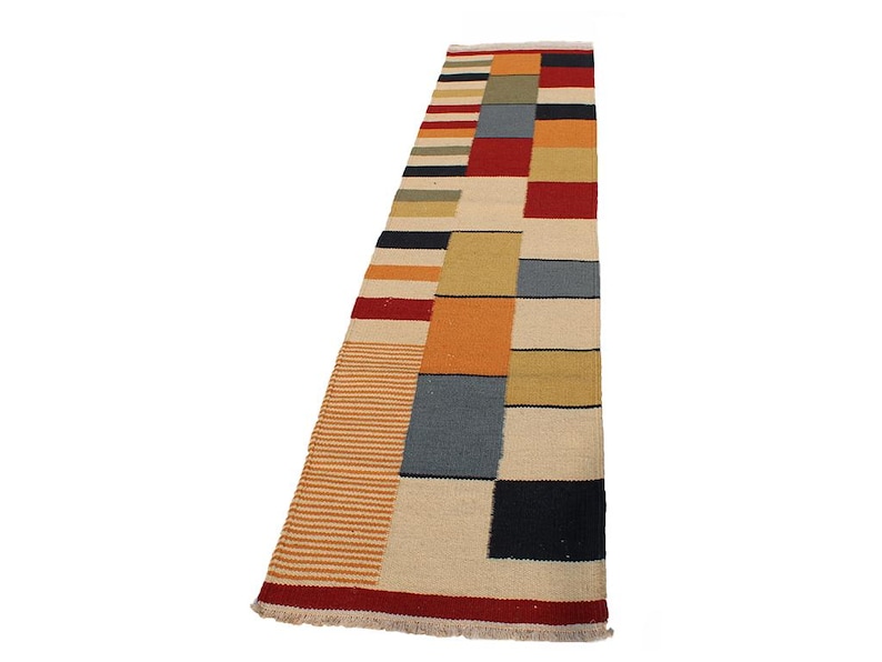 May include: A rectangular rug with a geometric pattern. The rug features a combination of colors, including beige, red, orange, yellow, blue, and black. The design includes stripes and rectangular blocks. The rug has a fringed edge.