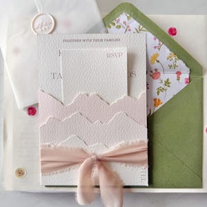 Peut inclure: Ensemble de faire-part de mariage avec des cartes blanches texturées et un motif rose en forme de montagne. Comprend une carte RSVP, une enveloppe verte avec une doublure florale et un ruban blush. Le texte "TOGETHER WITH THEIR FAMILIES" est visible.