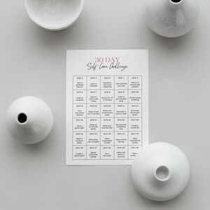 May include: A printable 30-day self-care challenge with a grid of daily activities. The grid is printed on white paper and has a pink title. The image also shows four white ceramic vases.