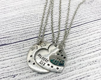May include: Three silver-toned necklaces with heart-shaped pendants. The pendants are engraved with the words "Big Sis", "Middle Sis", and "Little Sis". The necklaces are displayed on a light wood-grain surface.