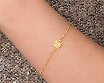 May include: A gold chain bracelet with a small square charm that has the word "Hamsa" engraved on it.