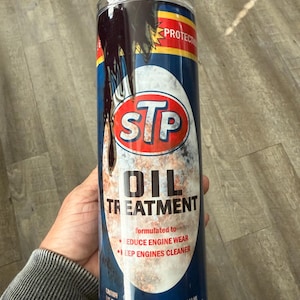 May include: A cylindrical tumbler featuring the STP Oil Treatment logo. The design includes a red and white STP logo, with the words "OIL TREATMENT" in bold letters. The tumbler is blue with black drips and the text "formulated to reduce engine wear" and "keep engines cleaner".