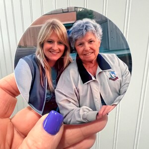 May include: A round photo of two women, one with blonde hair and one with gray hair, smiling at the camera. The woman with blonde hair is wearing a blue and gray jacket and the woman with gray hair is wearing a white jacket with a blue collar and a patch on the left side.