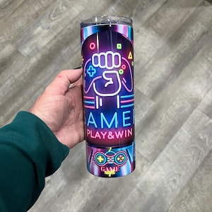 May include: A stainless steel tumbler with a neon pink, blue, and yellow gamer design. The tumbler features a fist holding a video game controller with the words "Gamer Play & Win" and "Game Night".