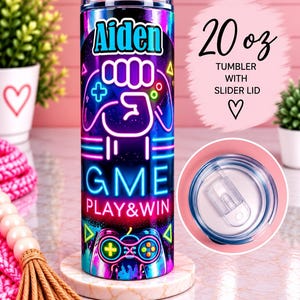 May include: A 20 oz tumbler with a slider lid, featuring a neon-style graphic print with the name Aiden, a fist icon, game controllers, and the text GME PLAY&WIN. The design uses vibrant blue, purple, and pink colors on a dark background.