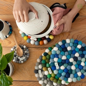 Può includere: Una varietà di sottopentola e sottobicchieri colorati a forma di palline di feltro sono esposti su una superficie di legno. Una pentola di ceramica bianca macchiata è tenuta sopra un sottopentola con un arcobaleno di palline di feltro. Sono visibili anche un cucchiaio di legno e una tazza di caffè.