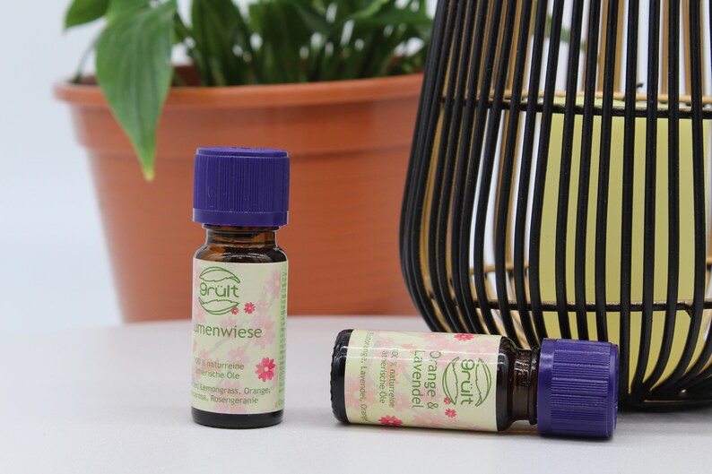 May include: Two small glass bottles with purple lids and labels that say "Gr&uuml;lt" and "Orange & Lavendel" in pink and green lettering. The bottles are sitting on a white surface.