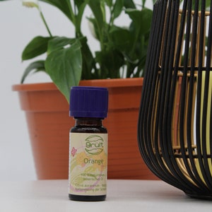 May include: A small, dark brown glass bottle with a purple cap. The bottle is labeled "Grut" and "Orange" and contains 100% pure essential oil. The bottle is sitting on a white surface next to a potted plant and a black and gold decorative object.