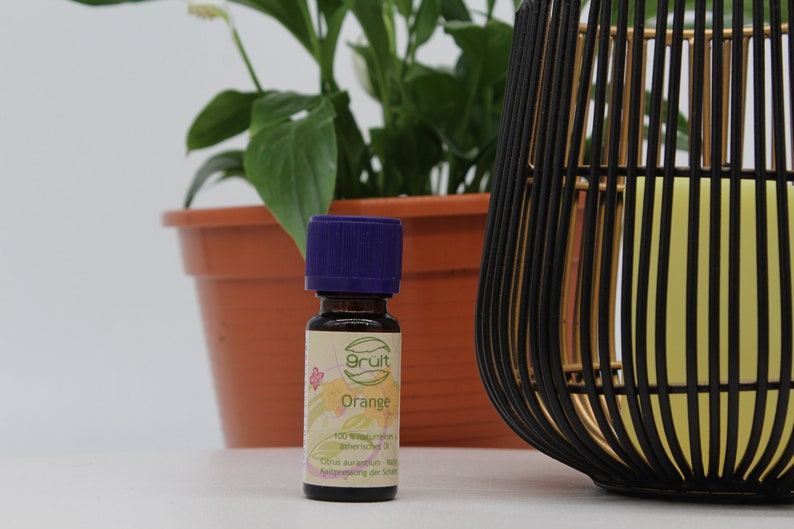 May include: A small, dark brown glass bottle with a purple cap. The bottle is labeled "Grut" and "Orange" and contains 100% pure essential oil. The bottle is sitting on a white surface next to a potted plant and a black and gold decorative object.