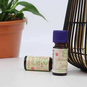 May include: Two small glass bottles of essential oil with purple lids. The bottles are labeled "Orange & Lavender" and "Blumenwiese". The bottles are sitting on a white surface next to a black and gold decorative cage.