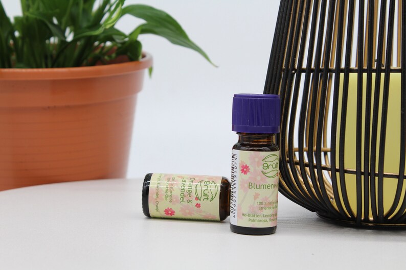 May include: Two small glass bottles of essential oil with purple lids. The bottles are labeled "Orange & Lavender" and "Blumenwiese". The bottles are sitting on a white surface next to a black and gold decorative cage.