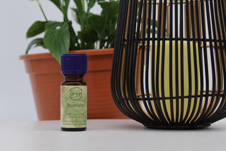 May include: A small brown glass bottle with a purple cap. The bottle is labeled "Rosmarin" and contains 100% natural rosemary essential oil. The bottle is sitting on a white surface next to a potted plant and a black and gold decorative lantern.