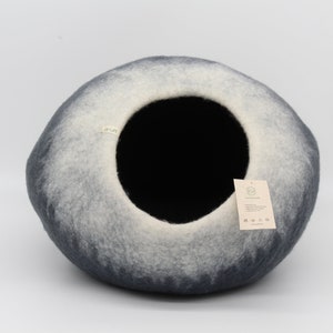 Może przedstawiać: Szare i białe filcowe legowisko dla kota w kształcie kopuły z czarnym otworem. Legowisko ma przyczepioną etykietę z napisem "Zrobione z miłością".