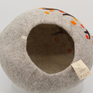 Może przedstawiać: Okrągłe legowisko dla kota z filcu w kolorze szarym z okrągłym otworem. Legowisko ma brązowo-pomarańczowy wz&oacute;r z małymi k&oacute;łkami na zewnątrz. Legowisko ma etykietę z napisem "Sprout".