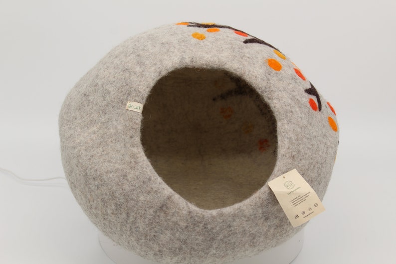 Może przedstawiać: Okrągłe legowisko dla kota z filcu w kolorze szarym z okrągłym otworem. Legowisko ma brązowo-pomarańczowy wz&oacute;r z małymi k&oacute;łkami na zewnątrz. Legowisko ma etykietę z napisem "Sprout".