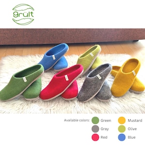 Op de afbeelding: Een selectie van wollen pantoffels in verschillende kleuren, waaronder groen, grijs, rood, mosterd, olijf en blauw. De pantoffels liggen op een wit, pluche oppervlak. De tekst "grült" is zichtbaar in de linkerbovenhoek van de afbeelding.